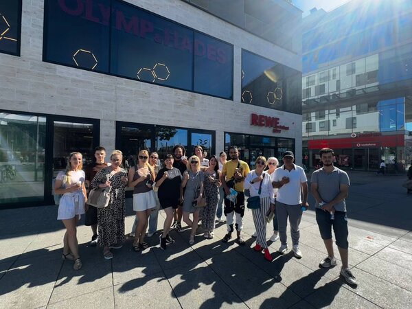 Eine Gruppe von etwa 18 jungen Erwachsenen steht bei sonnigem Wetter vor dem Einkaufszentrum Olymp & Hades in Leipzig. Im Hintergrund sind Schilder von REWE und Vodafone zu sehen. Die Gruppe hält Eis in der Hand und lächelt in die Kamera.
