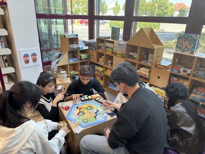 Eine Gruppe Jugendlicher und Kinder sitzt an einem Tisch und spielt das Brettspiel „Die Siedler von Catan“. Im Hintergrund stehen Regale voller Gesellschaftsspiele. Der Raum ist hell mit großen Fenstern, durch die man nach draußen sieht.