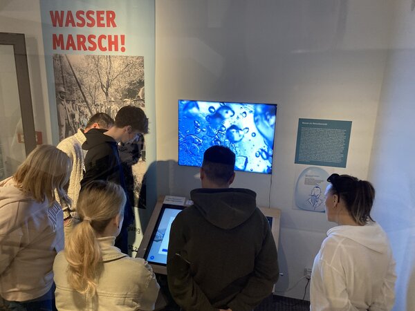 Exkursion ins Stadtmuseum Jena Eine Gruppe von Menschen steht vor einer interaktiven Ausstellung mit dem Thema Wasser. Sie schauen auf einen Bildschirm mit einer Nahaufnahme von Wassertropfen. Im Hintergrund ist ein Plakat mit der Aufschrift „WASSER MARSCH!“ zu sehen.