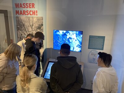Eine Gruppe von Menschen steht vor einer interaktiven Ausstellung mit dem Thema Wasser. Sie schauen auf einen Bildschirm mit einer Nahaufnahme von Wassertropfen. Im Hintergrund ist ein Plakat mit der Aufschrift „WASSER MARSCH!“ zu sehen.
