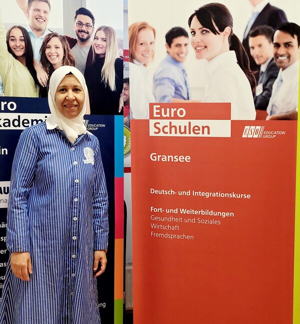 Neue Deutschdozentin Eine Frau mit einem langen blau-weißen Kleid und einen Kopftuch steht neben dem Roll-up der Euro-Schulen.