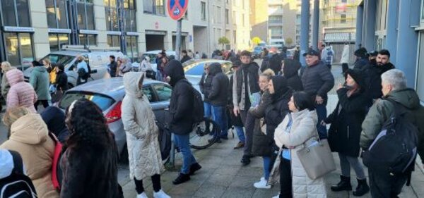 Eine große Gruppe von Menschen steht vor einem Gebäude, das mit 'Euro Akademie' beschriftet ist. Die Personen tragen warme Kleidung und scheinen sich in einer Wartesituation zu befinden. Autos und Fahrräder sind im Hintergrund zu sehen, ebenso wie ein Gehweg und Straßenschilder.