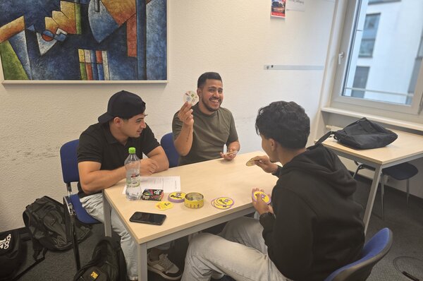 Spielen, Sprechen, Spaß haben – Kreative Gesprächsanlässe im Jugendintegrationskurs Drei junge Männer spielen ein Kartenspiel an einem Tisch. Einer hält lachend eine Karte hoch, während die anderen aufmerksam zuschauen. Auf dem Tisch liegen Karten und eine Getränkeflasche.