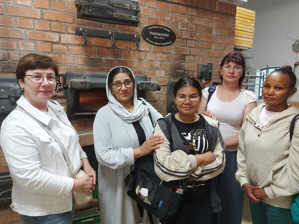 Praxis trifft Sprache - Hotel und Gastronomie Fünf Frauen stehen vor einer Backofenanlage mit Ziegelwand und blicken in die Kamera. Die Gruppe ist Teil einer Besichtigung oder eines Ausflugs.