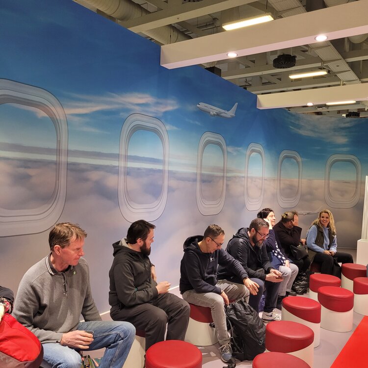 „BO auf Tour – Unternehmen laden ein“ auf der ITB 2025 Menschen sitzen auf runden, roten Hockern in einer Messe-Lounge, die einem Flugzeuginterieur mit Fenstern über den Wolken nachempfunden ist. Ein großes Wandbild zeigt einen Himmel mit einem fliegenden Flugzeug. Die Personen unterhalten sich oder schauen auf ihre Handys.