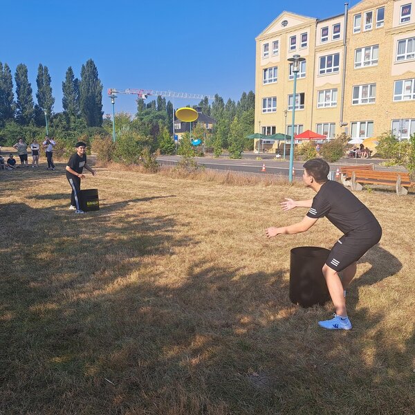 Sportfest – mal anders Zwei junge Männer werfen eine Frisbeescheibe, im Hintergrund stehen ein paar Zuschauer*innen. Auch das Schulgebäude ist im Hintergrund zu sehen.