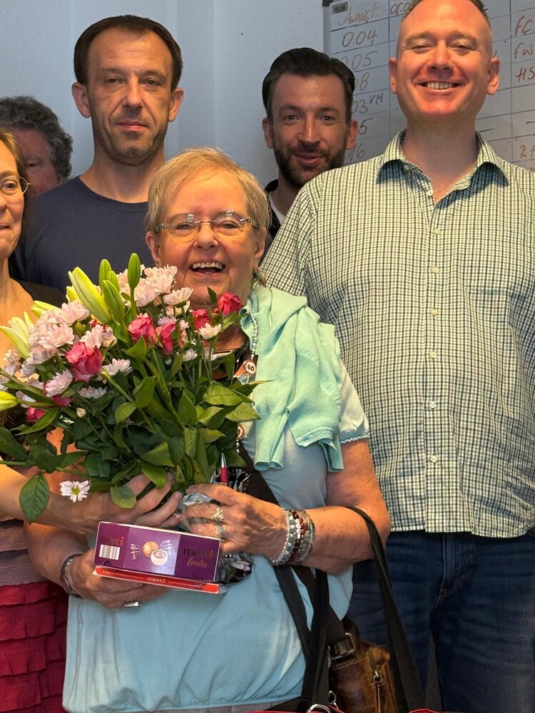 Nahaufnahme einer älteren Frau, die einen Blumenstrauß und eine Schachtel Pralinen hält. Sie steht lächelnd vor mehreren Personen, die sie umgeben. Im Hintergrund ist ein beschriftetes Whiteboard sichtbar.