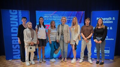 Vor einer blauen Wand mit digitalen Roll-ups der Euro Akademie steht in einem Raum eine Gruppe jüngerer und älterer Frauen sowie ein junger Mann.