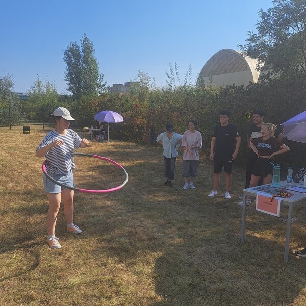 Sportfest – mal anders Eine Schülerin balanciert auf einer Wiese einen Hula Hoop-Reifen um ihre Taille, weitere Schüler*innen schauen ihr dabei zu.