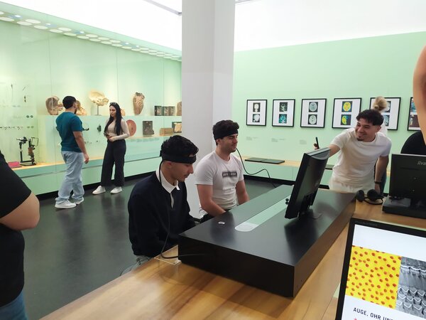 Sonniger Exkursionstag der Euro-Schulen Dresden: Spannende Einblicke in Wissenschaft und Kultur In einem interaktiven Museum sitzen zwei junge Männer mit Stirnbändern vor einem Bildschirm, während eine weitere Person lachend auf das Display zeigt.