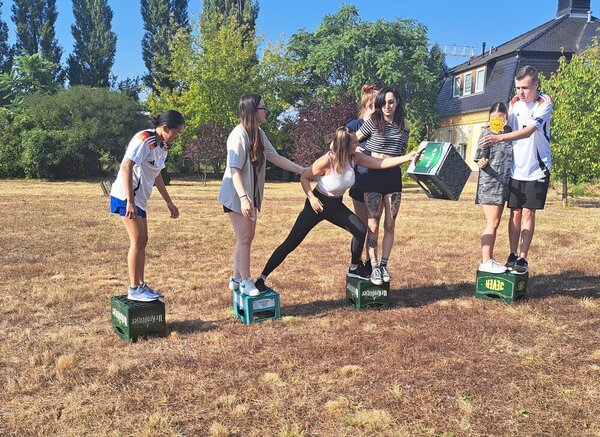 Sportfest – mal anders Mehrere Schülerinnen und Schüler stehen auf vier umgedrehten Bierkästen, teilweise befinden sich zwei Personen zusammen auf einem Kasten. Eine Schülerin wechselt gerade von einem Kasten zum nächsten und übergibt gerade einen Bierkasten an ein Teammitglied auf dem letzten Kasten.
