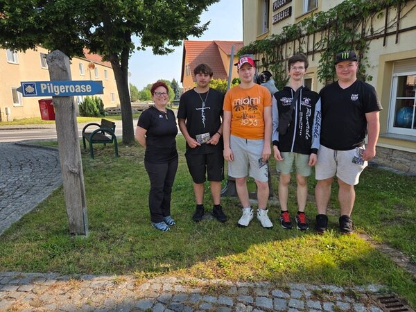 Pilgern mit Perspektive Die vier männlichen Auszubildenden zu Fachlageristen posieren, gemeinsam mit ihrer Lehrerin, für ein Gruppenfoto an einem Schild, auf dem Pilgeroase steht.