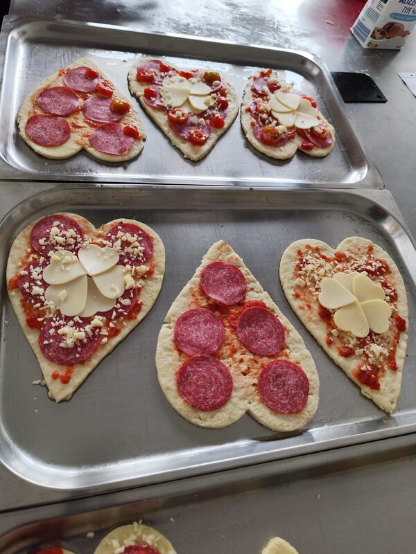 Praxisnahe Berufsorientierung: BRAFO im Strukturelement I Mehrere ungebackene, herzförmige Pizzen auf einem Backblech. Sie sind mit Salami, Käse und Tomaten belegt, einige mit dekorativen Käsestücken in Blumenform.