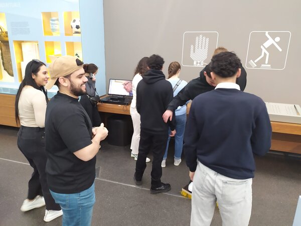 Sonniger Exkursionstag der Euro-Schulen Dresden: Spannende Einblicke in Wissenschaft und Kultur Besucher eines Museums stehen vor einer interaktiven Wand mit verschiedenen Symbolen und versuchen, ein Experiment mit einem Paar Bürstenschuhe.