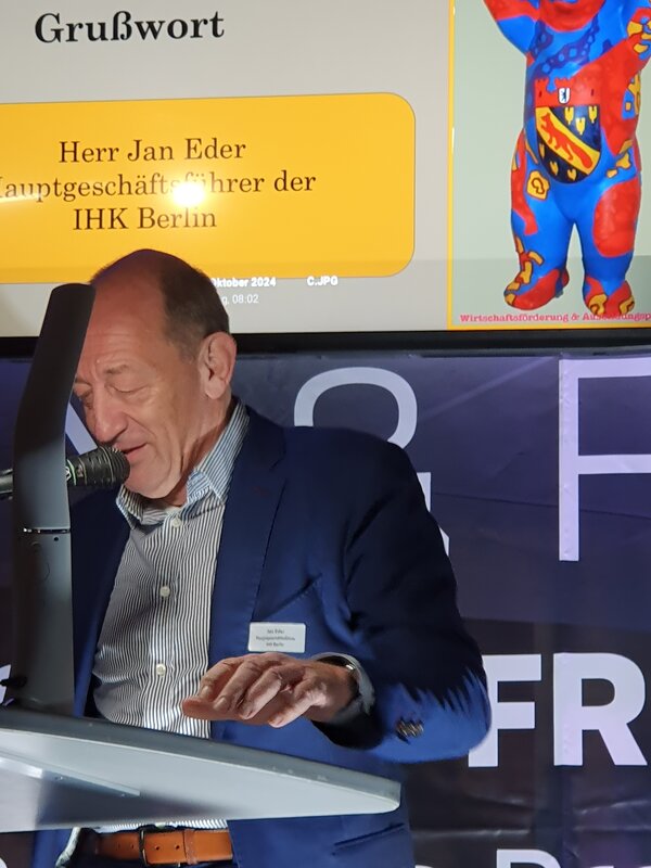 Reinickendorfer Ausbildungsbuddy geht an Netzwerkpartner der Euro-Schulen Berlin Ein Bühne mit grauem Rednerpult, an dem ein älterer Mann steht, und ein Bildschirm im Hintergrund mit der Aufschrift Ausbildungsbuddy 2024.