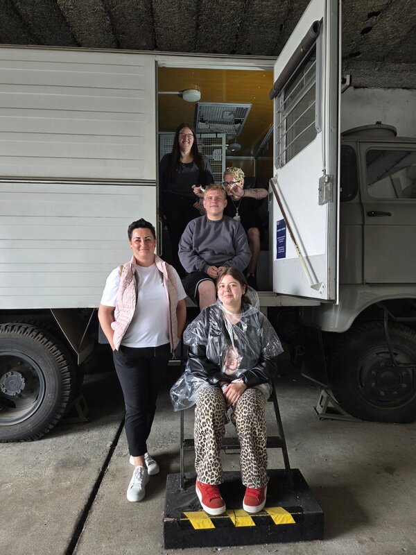 Exkursion zur Gedenkstätte Bautzen Fünf junge Menschen posieren vor einem weißen LKW mit offener Tür in einer Halle. Zwei Personen sitzen auf der Treppe zum Fahrzeug, eine Person sitzt vorne auf einem Hocker und trägt einen durchsichtigen Regencape. Zwei weitere Personen stehen seitlich und lächeln in die Kamera.