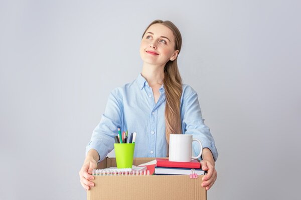 Eine junge Frau mit blonden Haaren, die zu einem Zopf nach hinten gebunden sind, schaut optimistisch und lächelnd nach hoben und hält einen geöffneten Karton mit Schreibwaren in den Händen.
