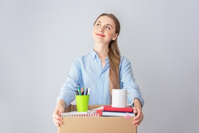 Optimistisch Eine junge Frau mit blonden Haaren, die zu einem Zopf nach hinten gebunden sind, schaut optimistisch und lächelnd nach hoben und hält einen geöffneten Karton mit Schreibwaren in den Händen.