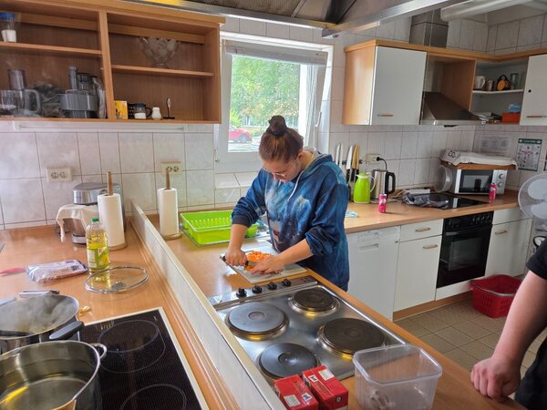 Lirum larum Löffelstiel Eine Person steht an einem Küchenarbeitsbereich und schneidet Karotten auf einem Schneidebrett. Die Küche ist mit verschiedenen Küchengeräten und Utensilien ausgestattet, darunter ein Herd, ein Kühlschrank, und mehrere Schränke. Es gibt auch verschiedene Küchenutensilien wie eine Flasche Öl, Gewürze, und Küchenwerkzeuge auf der Arbeitsplatte. Eine weitere Person ist teilweise sichtbar und steht in der Nähe.