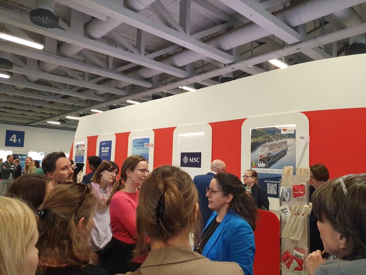 „BO auf Tour – Unternehmen laden ein“ auf der ITB 2025 Eine Gruppe von Menschen steht vor einem Messestand mit rot-weißer Gestaltung und Infotafeln von Unternehmen wie MSC und AIDA. Eine Frau im blauen Blazer spricht zu den Anwesenden. Der Stand scheint sich auf die Kreuzfahrt- oder Tourismusbranche zu beziehen.