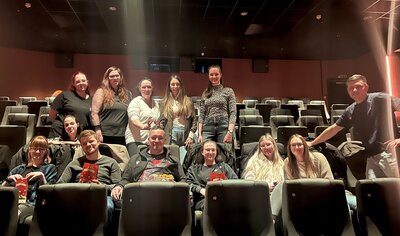 Die Auszubildenden im Kino Ein dunkler Kinosaal mit Sesseln. In den Reihen stehen und sitzen 13 junge Menschen und blicken lächelnd in die Kamera.