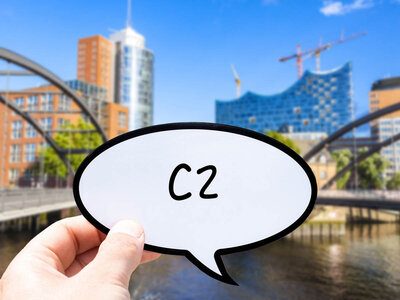 Eine Hand hält eine Sprechblase mit dem Text 'C2'. Im Hintergrund ist eine Stadtlandschaft mit modernen Gebäuden zu sehen, darunter ein markantes blaues Glasgebäude mit einem wellenförmigen Dach und Kräne in der Ferne. Im Vordergrund befindet sich eine Brücke über Wasser.