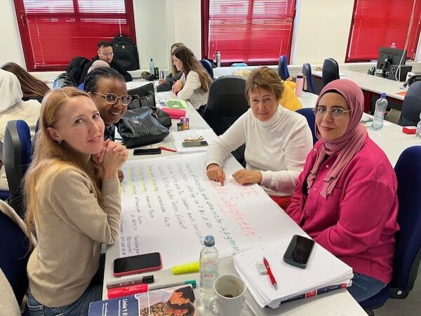 Erfolgreiche Gruppenarbeit im Berufssprachkurs Vier Teilnehmende des Berufssprachkurses sitzen an ihrer Projektarbeit und blicken in die Kamera.