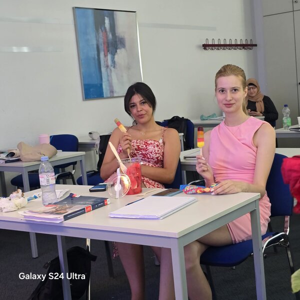 Zwei junge Frauen sitzen in einem Klassenzimmer an einem Tisch und genießen ein Eis am Stiel. Auf dem Tisch liegen Hefte, Stifte und weitere Lernmaterialien. Im Hintergrund sind weitere Tische und ein abstraktes Gemälde an der Wand zu sehen.