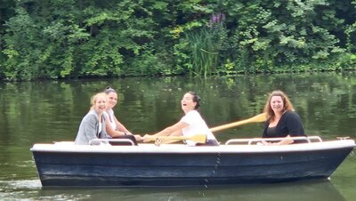 Vier Frauen sitzen in einem Boot auf einem See und lachen. Sie halten Paddel in den Händen und genießen die Zeit auf dem Wasser. Im Hintergrund sind Bäume zu sehen.