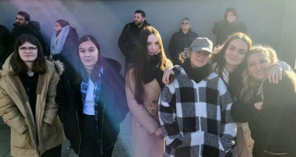 Eine Gruppe von sechs jungen Frauen steht nebeneinander draußen in einer Menschenmenge. Sie tragen warme Kleidung wie Mäntel und Jacken, passend für kühles Wetter. Einige der Frauen haben den Arm um ihre Freundinnen gelegt. Im Hintergrund stehen weitere Personen, die ebenfalls warme Kleidung tragen und teilweise von Sonnenlicht beschienen werden.