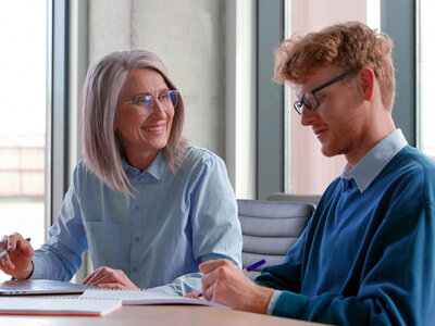 Eine ältere Frau mit grauen Haaren und Brille sitzt an einem Tisch und unterhält sich lächelnd mit einem jungen Mann mit roten Haaren und Brille. Beide tragen Hemden und scheinen in einem Büro oder Besprechungsraum zu sein. Die Frau hält einen Stift in der Hand und hat einen Laptop vor sich, während der Mann ein Notizbuch vor sich hat.