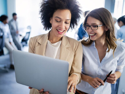 Zwei lächelnde Frauen in einem Büroumfeld, die gemeinsam auf einen Laptop schauen. Eine der Frauen trägt eine Brille und hält einen Stift, während die andere eine beige Jacke trägt. Im Hintergrund sind weitere Personen in einem hellen, modernen Büro zu sehen.