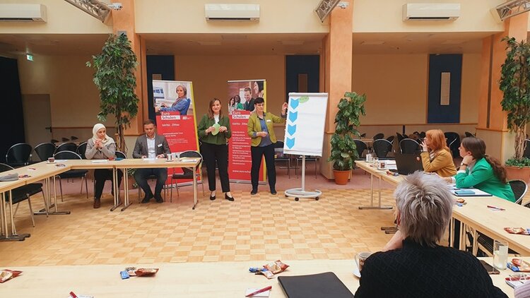 Gelungene Auftaktberatung der Arbeitsmarktmentoren Teilnehmende sitzen an Tischen und schauen interessiert nach vorn zu den zwei weiblichen Teilnehmenden, die an einem Flipchart Ergebnisse präsentieren.