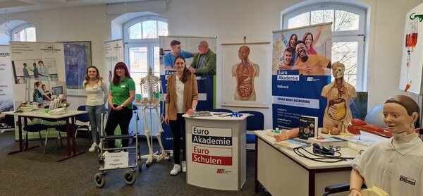 Hausmesse in Pößneck Ein Blick auf die gleiche Ausstellungsfläche aus einer leicht anderen Perspektive. Im Vordergrund ein Anatomie-Modell und ein Rollator mit dem Schild „Werde mit UNS Pflegeprofi“. Freundliche Standbetreuerinnen stehen bereit.