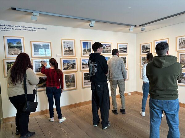 Exkursion ins Stadtmuseum Jena Eine Gruppe von Menschen betrachtet eine Fotoausstellung mit gerahmten Bildern von Brunnen und Wasserquellen. Ein Zitat an der Wand von Demosthenes warnt vor der Übernutzung von Wasserquellen: „Auch Quellen und Brunnen versiegen, wenn man zu oft und zu viel aus ihnen schöpft.“