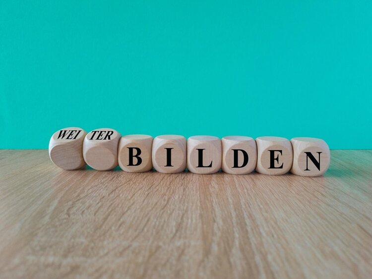 Auf hölzernen Würfeln stehen die Buchstaben, die das Wort Weiterbildung bilden. Die ersten Würfel, auf denen Weiter steht, sind leicht nach hinten geneigt, sodass das Wort Bildung im Fokus steht. Der Hintergrund ist türkisblau.