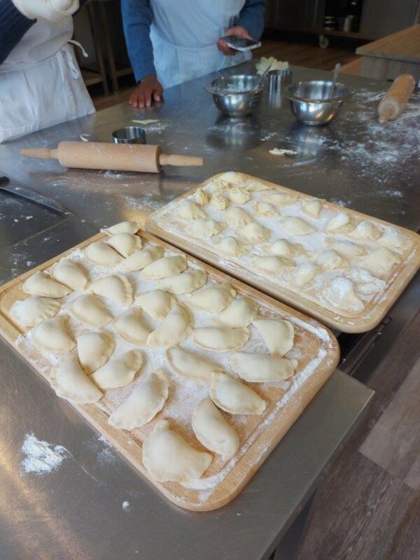 Praxis trifft Sprache - Hotel und Gastronomie Frisch zubereitete Teigtaschen liegen bemehlt auf zwei Holzbrettchen, bereit zum Kochen. Im Hintergrund liegen Nudelholz, Metallschüsseln und weitere Backutensilien auf einem Edelstahltisch.