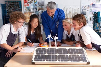 Ausbilder erklärt  Ein Ausbilder erklärt seinen Auszubildenden etwas über Windenergie und Solarpaneele in einer Berufsschule. Die weiblichen und männlichen Azubis beugen sich interessiert und gut gelaunt über das aufgebaute Model.