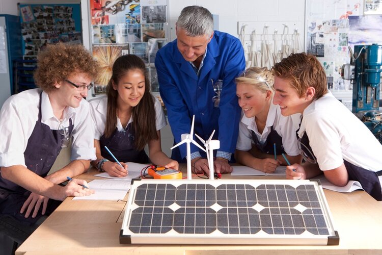 Ein Ausbilder erklärt seinen Auszubildenden etwas über Windenergie und Solarpaneele in einer Berufsschule. Die weiblichen und männlichen Azubis beugen sich interessiert und gut gelaunt über das aufgebaute Model.