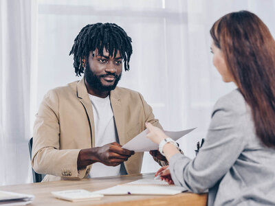 Beratung und Betreuung anerkannter Flüchtlinge in Thüringen (BeraFiT) Ein Mann mit Dreadlocks und einem hellbraunen Blazer sitzt an einem Schreibtisch und reicht einer Frau in einem grauen Anzug ein Dokument. Beide wirken professionell und sind in ein Gespräch vertieft. Auf dem Tisch liegen verschiedene Unterlagen und ein Laptop.