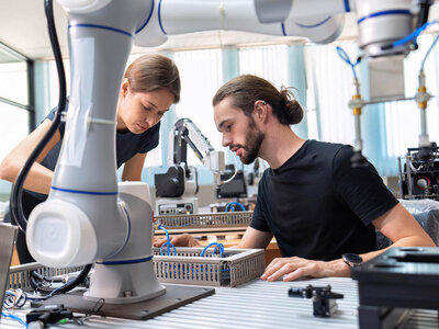 Zwei Personen in einem technischen Labor arbeiten an einem Projekt mit einem Roboterarm. Die Frau mit kurzen Haaren und schwarzem T-Shirt beobachtet den Roboterarm, während der Mann mit langem Haar und Bart an einem Gerät auf dem Tisch arbeitet. Im Hintergrund sind verschiedene technische Geräte und Maschinen zu sehen, die auf eine industrielle oder Forschungsarbeit hinweisen.