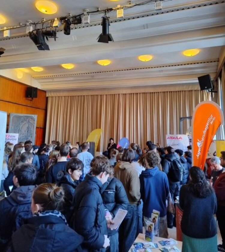 Zukunftstag in Berlin-Reinickendorf Ein großer Raum mit vielen jungen Menschen, die sich an verschiedenen Infoständen über Bildungs- und Berufsmöglichkeiten informieren. Auf einer kleinen Bühne spricht eine Person vor dem Publikum, während bunte Banner und Informationsmaterialien die Veranstaltung begleiten.