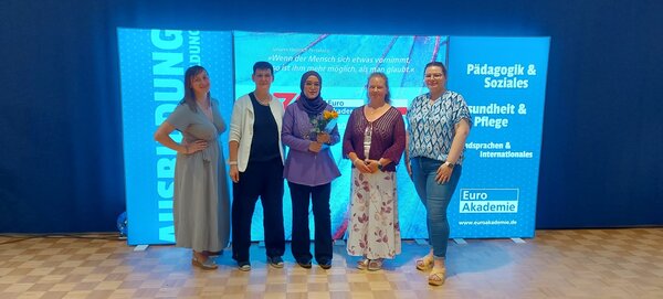 Erfolgsstory: Ein Meilenstein voller Stolz und Hoffnung Fünf Frauen stehen nebeneinander auf einem Parkettboden vor einem großen, beleuchteten Hintergrund mit der Aufschrift "Euro Akademie" und den Bereichen "Pädagogik & Soziales", "Gesundheit & Pflege" sowie "Fremdsprachen & Internationales". Die Frau in der Mitte hält einen Blumenstrauß. Alle Personen blicken in die Kamera und lächeln.