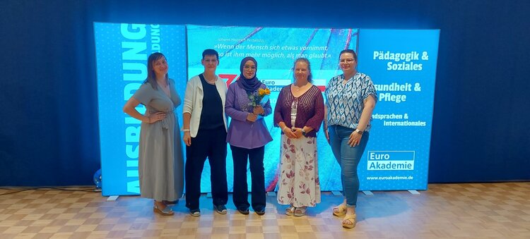 Erfolgsstory: Ein Meilenstein voller Stolz und Hoffnung Fünf Frauen stehen nebeneinander auf einem Parkettboden vor einem großen, beleuchteten Hintergrund mit der Aufschrift "Euro Akademie" und den Bereichen "Pädagogik & Soziales", "Gesundheit & Pflege" sowie "Fremdsprachen & Internationales". Die Frau in der Mitte hält einen Blumenstrauß. Alle Personen blicken in die Kamera und lächeln.
