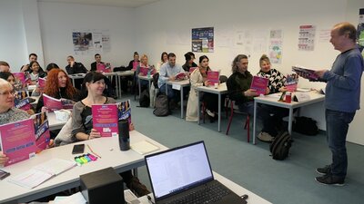 Teilnehmende sitzen gut gelaunt im Klassenraum an Tischen, alle mit aufgeklappten Informationsbroschüre in den Händen, der Lehrer begrüßt sie freundlich.