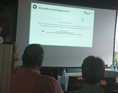Teilnehmende sitzen in einem Raum und schauen auf eine an die Wand projizierte PowerPoint-Präsentation.