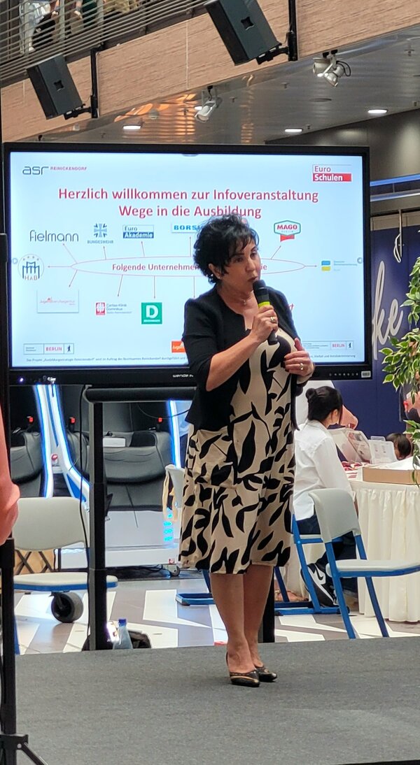 „Wege in die Ausbildung“ - Ein Tag voller Perspektiven für junge Menschen Die Bezirksbürgermeisterin Emine Demirbüken-Wegner begrüßt die Gäste der Veranstaltung, hinter ihr ein Bildschirm, au der alle beteiligten Unternehmen aufgeführt sind.