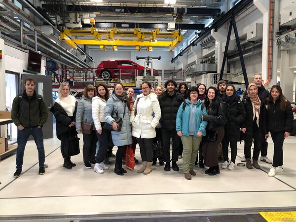 Sonniger Exkursionstag der Euro-Schulen Dresden: Spannende Einblicke in Wissenschaft und Kultur Eine große Gruppe von Personen steht in einer Werkshalle oder einem technischen Labor mit einem roten Auto im Hintergrund. Sie tragen Winterjacken und posieren für ein Gruppenfoto.