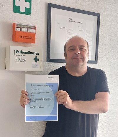 Ein Mann in schwarzem T-Shirt steht vor einer weißen Wand mit Erste-Hilfe Kasten und hält ein hellblaues Zertifikat in die Kamera.