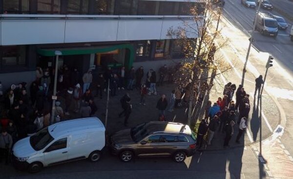 Eine große Menschenmenge steht in einer Schlange vor einem Gebäude. Die Schlange reicht bis zur Straßenecke, an der mehrere Autos und ein Baum zu sehen sind. Im Hintergrund ist eine stark befahrene Straße mit weiteren Fahrzeugen zu erkennen.